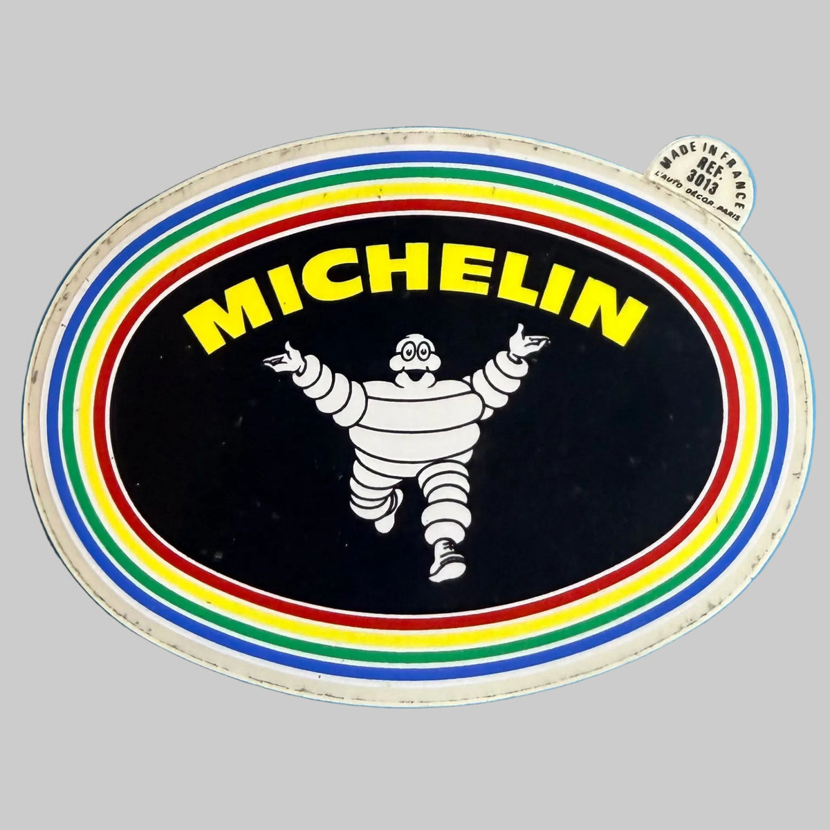 Autocollant Michelin vintage ovale avec Bibendum blanc, fond noir et bordures multicolores, fabrication française.