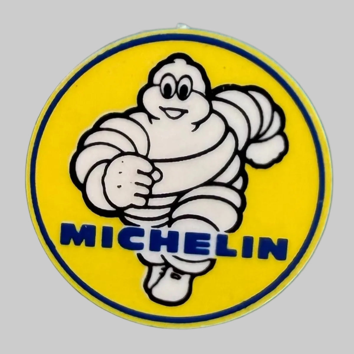 Badge Michelin Bibendum rond de 3 cm de diamètre, avec illustration du célèbre personnage en mouvement sur fond jaune et inscription MICHELIN bleue.