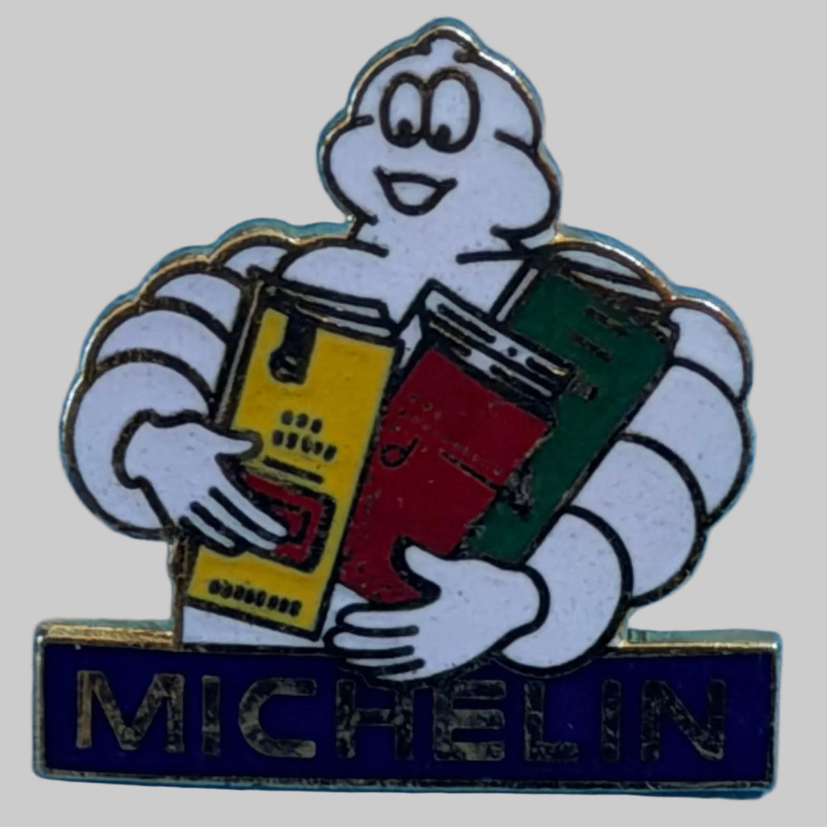 Pin's Michelin Bibendum tenant trois guides Michelin colorés, avec inscription Michelin sur fond bleu.