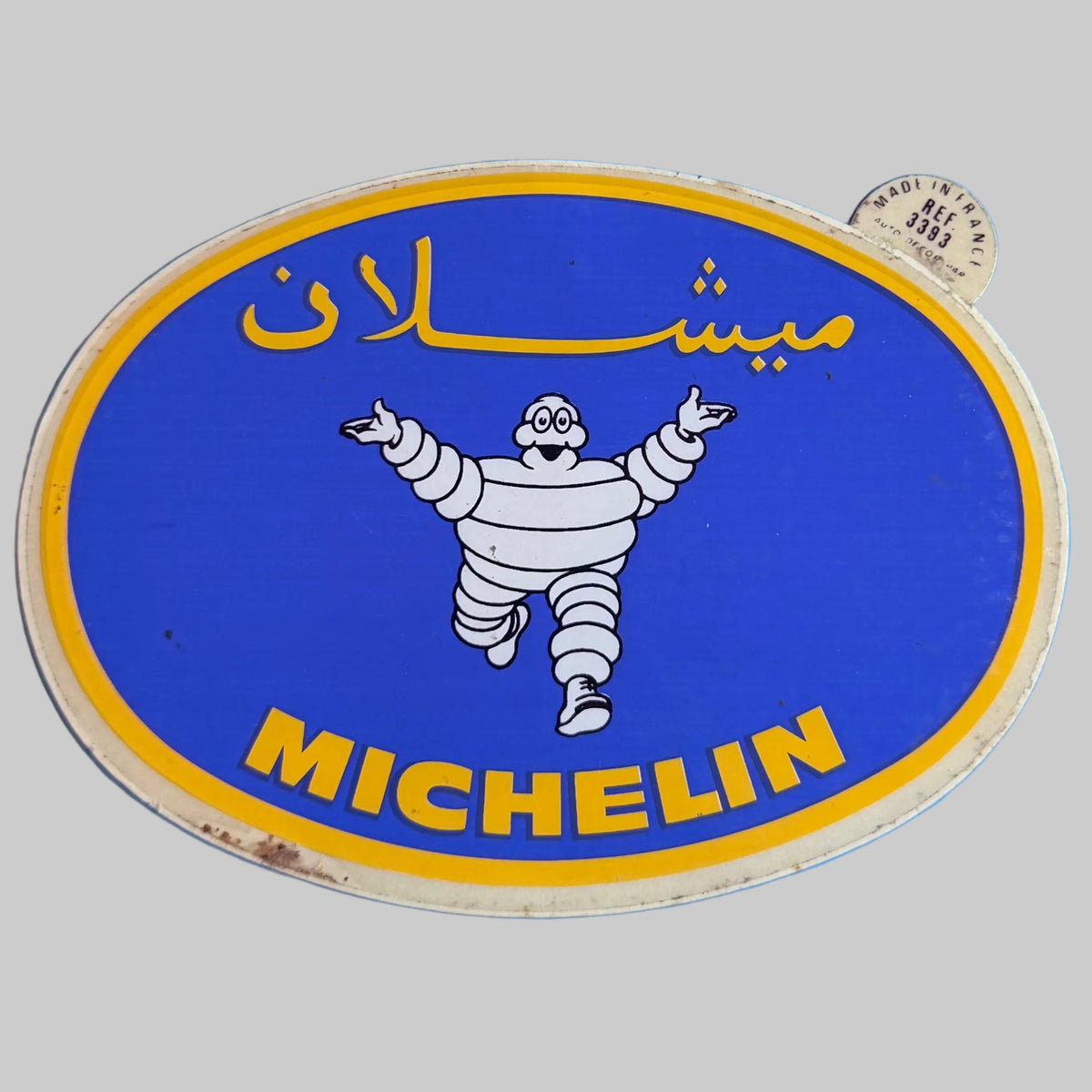 Autocollant Michelin ovale bleu et jaune avec Bibendum au centre, inscription arabe et logo Michelin.